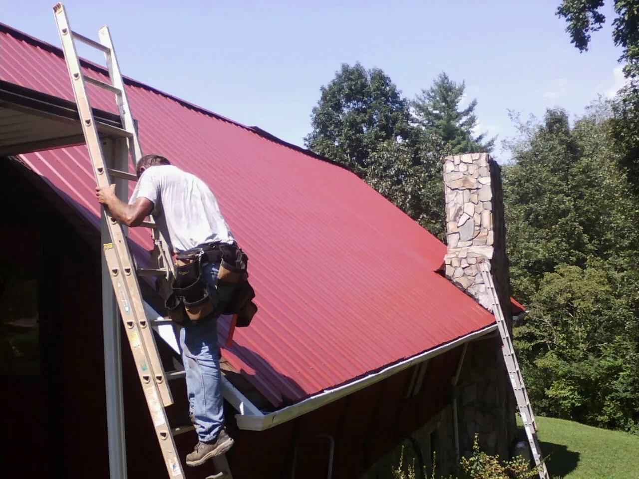 Roofer on a ladder beside a red metal roof and stone chimney — Soffit & Fascia Repair by Roofing Brothers in Milliken
